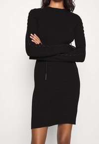 Robe noire en maille côtelée à manches longues, avec des détails texturés le long des manches et une silhouette ajustée, se terminant au-dessus du genou.