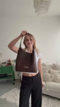 Brun syntetisk handväska med en avslappnad form, lång axelrem och en framträdande logotyp. Slät textur och minimalistisk design.