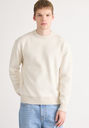 Pull à côtes crème avec un col rond et des manches longues, présentant une finition texturée. Associé à un jean en denim bleu clair.