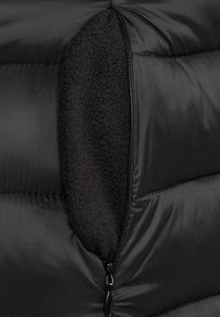 Détail d'une doudoune noire montrant une poche zippée doublée de doux polaire noir. Le tissu extérieur est lisse et brillant, avec des sections matelassées.