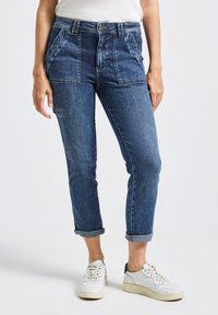 Hochtaillierte Jeanshose in Dunkelblau mit Fronttaschen und umgeschlagenen Säumen. Kombiniert mit weißen Sneakers. Der Stoff hat eine glatte Textur.