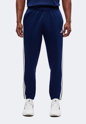 Άνδρας που φοράει σκούρο μπλε φόρμα Adidas με λευκές ρίγες και λευκά αθλητικά παπούτσια Adidas, στέκεται μπροστά σε μονόχρωμο φόντο.