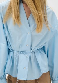 Light blue button-up blouse with a wrap design, cinched waist, and voluminous sleeves. Made of smooth fabric with a slight sheen.