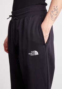 Black joggers made of soft fabric, featuring an elastic waistband with a drawstring and a white embroidered logo on the left thigh.