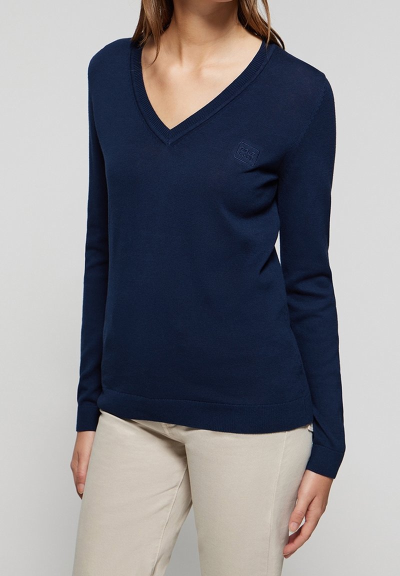 Femme portant un pull bleu marine à col en V à manches longues et un pantalon beige, debout devant un fond clair uni.