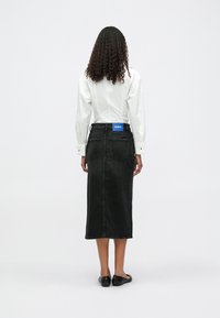 Femme aux cheveux bouclés portant une chemise blanche, une jupe en denim noire midi avec une étiquette Hugo et des ballerines noires, se tenant devant un fond uni.