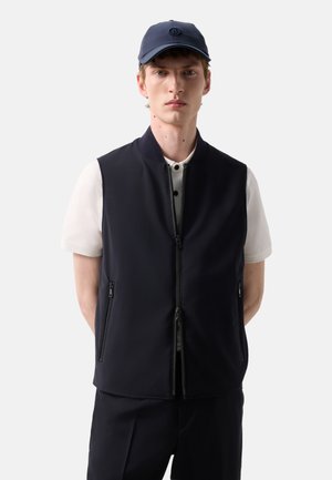 Młody mężczyzna w granatowej kamizelce zapinanej na zamek, na białej koszulce z krótkim rękawem oraz w pasującej granatowej czapce z daszkiem, stoi na jednolitym tle.