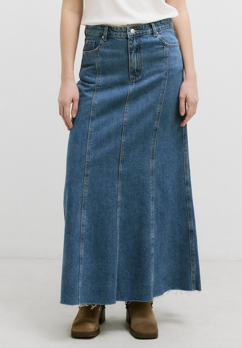 Femme portant une longue jupe en denim bleu avec un ourlet brut et des bottines marron, se tenant devant un fond blanc uni.