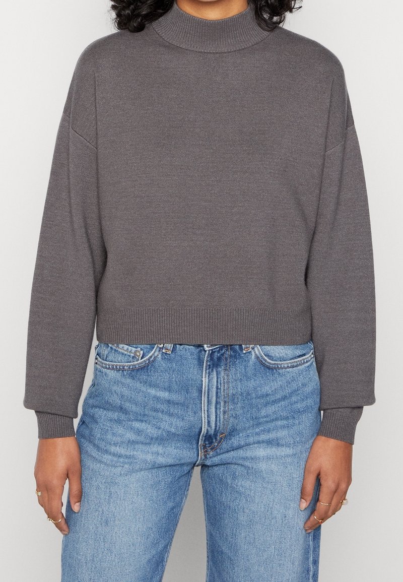 Personne portant un pull gris court à col roulé avec des manches longues et un jean bleu taille haute, debout devant un fond uni.