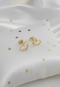 Guldhjärtlika stiftörhängen på en vit satinkudde, spridda med små guldhjärtformade konfetti. Slät, glänsande yta.