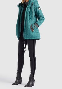 Femme portant une doudoune matelassée bleu sarcelle avec capuche, un pantalon slim noir avec une bande latérale, et des bottines à talons noires, debout les mains dans les poches.