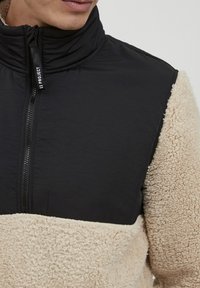 Black and beige fleece jacket with a high collar, zip closure, and textured fabric. The upper section is smooth, contrasting with the lower fleece.