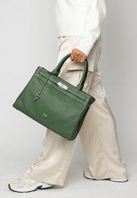 Sac à main en cuir vert avec surface texturée, poignées supérieures et ferrures argentées. Comprend un pendentif avec un "P". Porté avec un pantalon crème et des baskets.