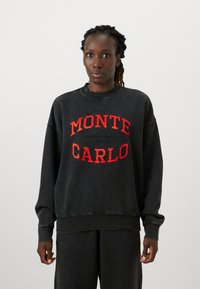 Mustat hupparit, joissa on rento istuvuus, ja joissa on punaisella brodeerattu teksti "MONTE CARLO." Kangas on pehmeä ja hihansuissa on ribbikuvio.