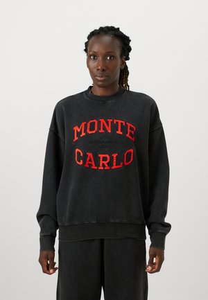 Sweatshirt noir avec une coupe décontractée, arborant le texte brodé rouge "MONTE CARLO". Le tissu a une texture douce et des poignets côtelés.