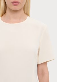 Haut à manches courtes de couleur crème avec un col arrondi. Tissu lisse avec une texture douce, détails de couture minimalistes.