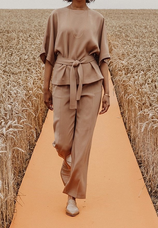 Person som går på en orange catwalk genom ett vetefält, klädd i en lös beige topp knuten i midjan och matchande vida byxor med beiga skor.