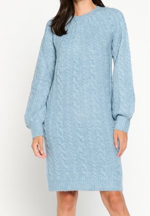 Kvinde iført en lyseblå, langærmet kabelstrikket sweaterkjole med ribkant ved ærmer og kant, stående mod en neutral baggrund.