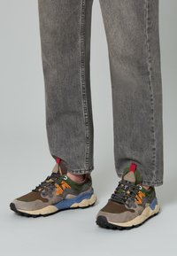 Mehrfarbige Sneakers, die eine Mischung aus braunen, grünen und blauen Materialien aufweisen, mit Wildlederstruktur und einer markanten Gummisohle mit ausgeprägten Stollen.