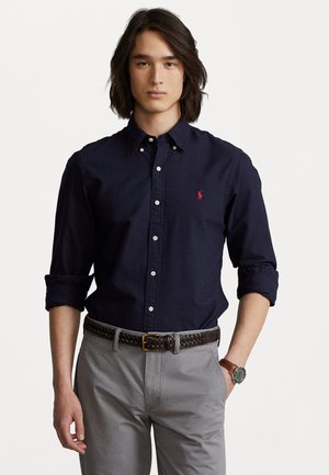 Camicia button-down blu in cotone con maniche lunghe arrotolate, caratterizzata da un piccolo logo ricamato rosso, abbinata a pantaloni grigi e una cintura intrecciata marrone.
