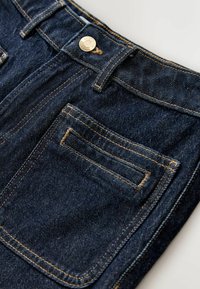 Mørkeblå denimjeans tæt på med en baglomme med orange syninger og en metalknap på linningen.