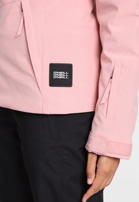 Nahaufnahme einer Person, die eine pinke Jacke mit einem schwarzen O'Neill-Logopatch und schwarzen Hosen trägt, wobei Ärmel- und Seitendetails zu sehen sind.