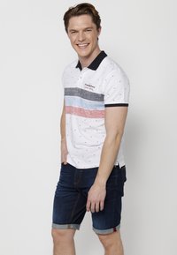 Polo majica v beli barvi z mornarsko modrim ovratnikom, s horizontalnimi črtami v črni, modri in rdeči barvi, okrašena z malimi geometrijskimi vzorci. Obuta z denim kratkimi hlačami.