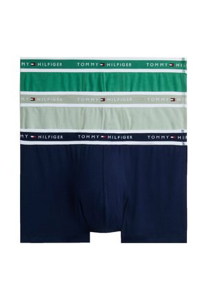 Hombre atlético de pie descalzo vistiendo bóxers azul marino con la cintura con la marca Tommy Hilfiger, mirando hacia la izquierda.