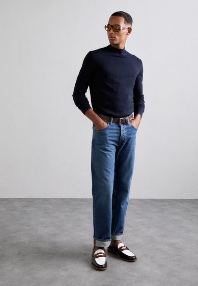 Turtleneck marine, jean bleu clair avec les poignets repliés, et mocassins marron et blanc. Des lunettes de soleil brillantes complètent la tenue.