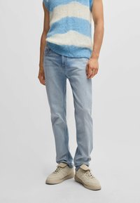 Jeans in denim azzurro chiaro con vestibilità dritta, caratterizzati da un classico design a cinque tasche e una leggera scoloritura; indossati con un gilet a righe blu e bianche senza maniche e sneakers beige.