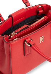 Κόκκινη δερμάτινη τσάντα χειρός με χρυσό λογότυπο TT, διπλές λαβές και μερικώς ανοιχτό επάνω φερμουάρ που αποκαλύπτει μαύρη εσωτερική επένδυση.