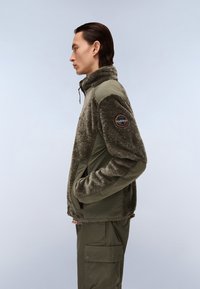 Vihreä fleece-takki, jossa on teksturoidu kangas, korkea kaulus ja vetoketjusuljenta. Ominaisuuksiin kuuluu kontrastiväriset kudotut paneelit ja pyöreä logo hihoissa.