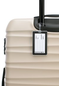 Maleta rígida beige con asa negra y un portaetiquetas de equipaje de plástico transparente adjunto con campos en blanco para información de contacto.