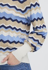 Pull tricoté à motif ondulé dans des nuances de bleu, beige et blanc. Poignets et bord inférieur côtelés, avec des points texturés créant de la profondeur.