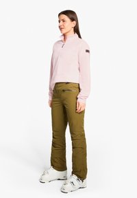 Helles rosa Fleece-Shirt mit Viertelreißverschluss, olivgrüne Skihosen mit Seitentasche, getragen mit weißen Skischuhen mit silberner Hardware.