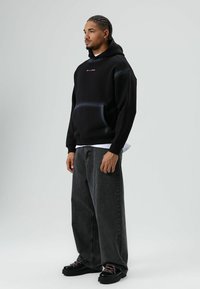 Zwarte hoodie met capuchon en een voorzak, voorzien van roze tekst. Draagt bijpassende donkergrijze jeans met wijde pijpen en zwarte veter schoenen.