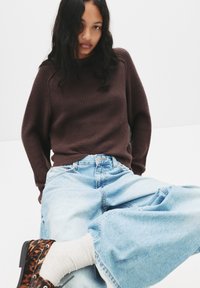 Brauner gerippter Pullover, hellblaue lockere Jeans, gemusterte Stiefeletten. Der Pullover hat einen hohen Kragen; die Jeans sind weit geschnitten und haben eine Jeansstruktur.