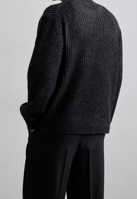 Pull en maille côtelée noire à coupe ample, avec un col rond et une texture subtile. Associé à un pantalon noir sur mesure.