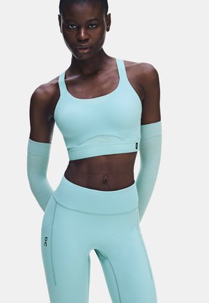 Frau steht frontal, trägt einen passenden hellblauen Sport-BH, Leggings und Armstulpen vor einem schlichten, hellen Hintergrund.