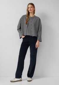 Grauer, verkürzter Pullover mit Rundhalsausschnitt und Knöpfen, kombiniert mit navyfarbener Hose. Das Modell trägt weiße Sneakers mit beigen Akzenten.