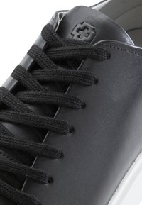 Černé kožené tenisky s plochými černými tkaničkami, hladkým povrchem a bílou podrážkou; zahrnují malé vyražené logo na jazyku.