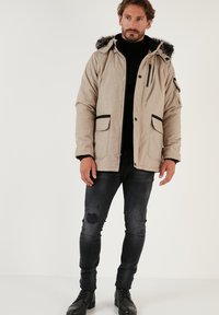 Beidzēts jaku ar bežīgu krāsu un vilnas apakšmali, melniem akcentiem, rāvējslēdzēja kabatu un divām priekšējām kabatām, valkāta virs melnā turtlenecka un nolietotām džinsiem.