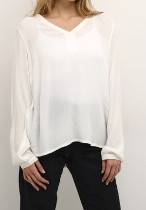Hvid bluse med lange ærmer, V-hals og struktureret stof; har et diskret forstykke og en løs pasform. Kombineret med sorte bukser.