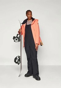 Snovborda tērps ar spilgtu korallkrāsas jakku, melnām kombinezona biksēm un melniem snovborda zābakiem. Tur rokās snovbordu ar stiprinājumiem.