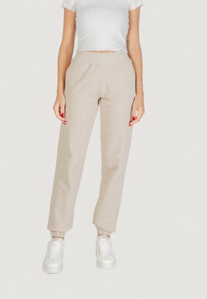 Femme portant un pantalon jogger beige à taille élastique, un t-shirt court côtelé blanc et des baskets blanches, debout devant un fond uni.