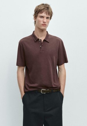 Jonge man die een bruin poloshirt met korte mouwen draagt, ingestopt in een zwarte broek met een gevlochten bruine riem, tegen een eenvoudige lichte achtergrond.