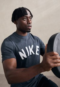 Pelēks Nike T-krekls ar baltu nolietotu logo, koncentrējoties uz indivīdu, kurš tur melnu svaru plati minimālisma vidē.