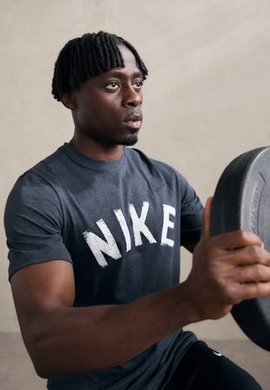 Gray Nike t-shirt with white distressed logo, focused on individual holding a black weight plate in a minimalist setting.