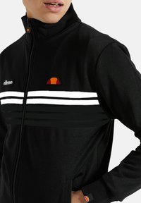 Zwarte zip-up jas met een hoge kraag, voorzien van witte horizontale strepen en een kleurrijke logo op de borst en mouw. Zachte materiaalkwaliteit.
