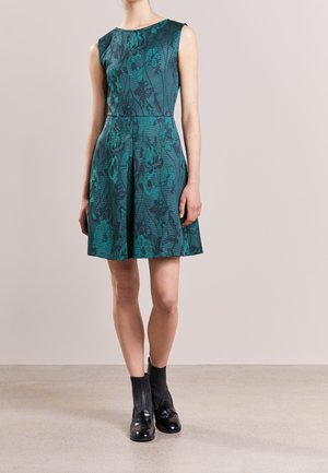 Mouwloze, knielange teal jurk met donker bloemmotief, gedragen met zwarte geribbelde sokken en zwarte glanzende loafers op een neutrale achtergrond.